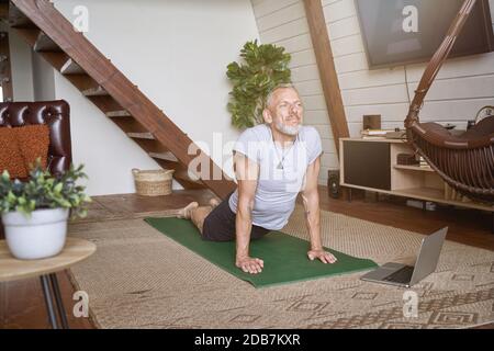 Sportlicher Mann mittleren Alters, der Yoga-Übungen im Leben macht Zimmer zu Hause, während Sie den Online-Kurs auf dem Laptop ansehen Stockfoto