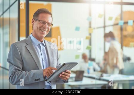 Fröhliche reife Geschäftsmann in klassischen tragen halten digitale Tablette und Lächelnd in der Kamera, während Sie im modernen Büro stehen Stockfoto