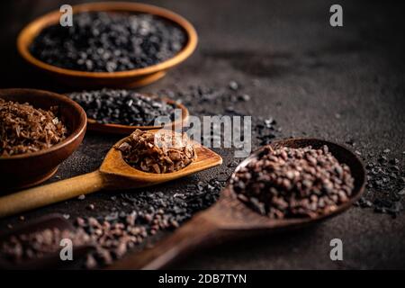 Verschiedene Gourmetsalze in Löffeln und Schüsseln auf dunklem Hintergrund Stockfoto