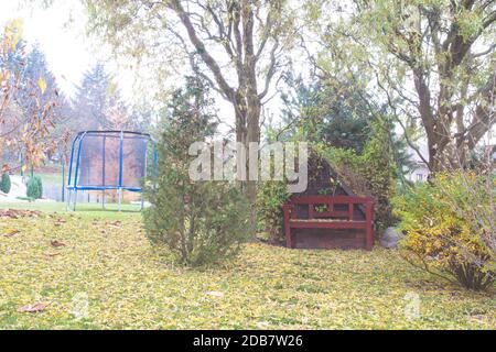 Bank im Garten in der Herbstsaison mit Blättern auf dem Boden. Trampolin im Garten. Bäume und Sträucher verloren Laub und sind bereit für den Winter. Stockfoto