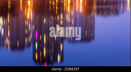 Silhouetten mehrstöckiger Häuser mit hellem Licht in den Fenstern spiegeln sich auf der blauen Wasseroberfläche im See. Große Stadt im Detail Stockfoto