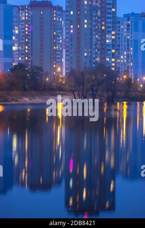 Silhouetten mehrstöckiger Häuser mit hellem Licht in den Fenstern spiegeln sich auf der blauen Wasseroberfläche im See. Große Stadt im Detail Stockfoto