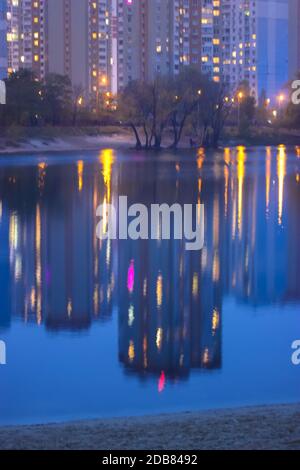 Silhouetten mehrstöckiger Häuser mit hellem Licht in den Fenstern spiegeln sich auf der blauen Wasseroberfläche im See. Große Stadt im Detail Stockfoto