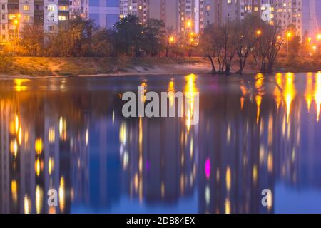 Silhouetten mehrstöckiger Häuser mit hellem Licht in den Fenstern spiegeln sich auf der blauen Wasseroberfläche im See. Große Stadt im Detail Stockfoto