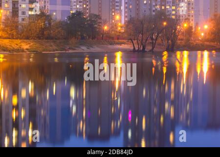 Silhouetten mehrstöckiger Häuser mit hellem Licht in den Fenstern spiegeln sich auf der blauen Wasseroberfläche im See. Promenade am See bei Nacht Stockfoto