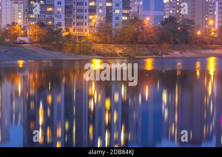 Silhouetten mehrstöckiger Häuser mit hellem Licht in den Fenstern spiegeln sich auf der blauen Wasseroberfläche im See. Promenade am See bei Nacht Stockfoto