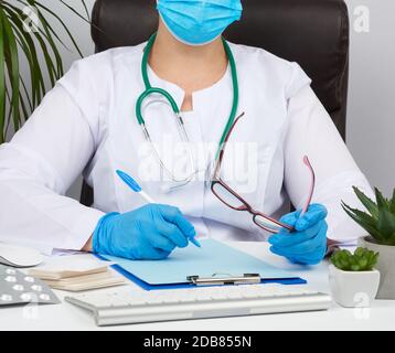 Junge Frau Doktor in einem weißen Mantel, sterile medizinische Handschuhe schreiben ein Rezept auf einer Form, Therapeut sitzt auf einem Stuhl an einem weißen Tisch, Arbeitsplatz Stockfoto