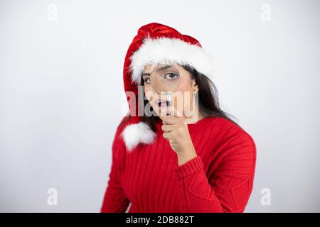 Junge schöne Frau trägt einen Weihnachtsmann Hut auf weißem Hintergrund Überrascht durch ein Vergrößerungsglas zu schauen Stockfoto