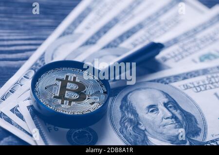 Bitcoin statt Lupe über dem Hintergrund von hundert-Dollar-Scheinen. Tonung in Blau. Das Konzept, Papiergeld durch virtuelles Geld zu ersetzen Stockfoto