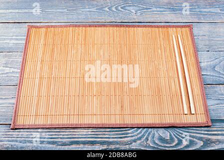 Zwei hölzerne Essstäbchen liegen auf dem Rand einer Bambusmatte, die auf einem Holztisch aus strukturierten Brettern sitzt. Stockfoto