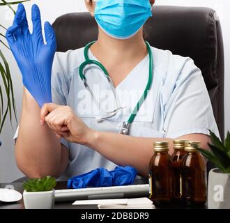 Frauenarzt in blauer Uniform, einmal-medizinische Maske sitzt in einem Stuhl am Tisch und legt ihm blaue sterile Latexhandschuhe, Doktor o, an Stockfoto