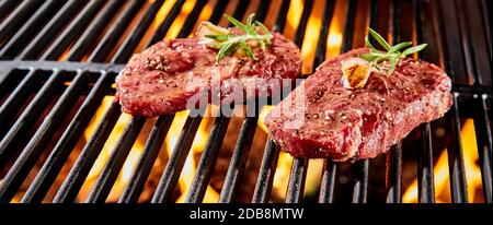 Zwei Portionen rohes Rinderfilet oder Rumpsteak, gewürzt mit Kräutern und Gewürzen, die über einem Grillfeuer in Nahaufnahme in einem Panorama-Banner grillen Stockfoto