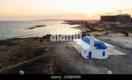 Blick auf den Sonnenuntergang an der Küste und die traditionelle weiße, gewaschene Kapelle mit blauen Türen am Strand von Agia Thekla, Ayia Napa, Famagusta, Zypern von A Stockfoto