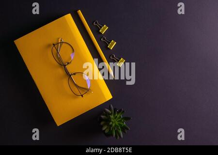 Gelber Bleistift, gelbes Notizbuch und Brille, Stifte, Clip-Papier auf schwarzem Hintergrund. Stockfoto