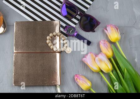 Ein goldenes Notizbuch mit Damenschuhen, Tulpen, Brillen und einer Uhr auf betonierten Hintergrund. Flet-Lay. Stockfoto