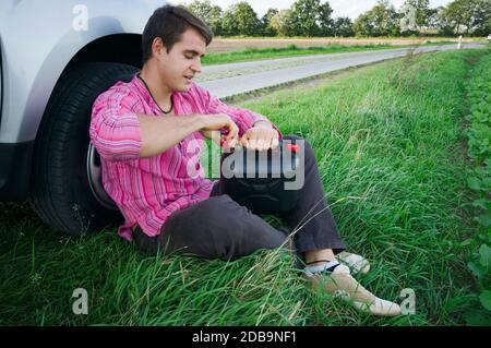 Seitliche Ganzkörperansicht eines jungen Mannes, der am Rand einer Landstraße im Gras auf dem Vorderreifen seines Autos sitzt und die Kappe des Kanisters öffnet. Stockfoto