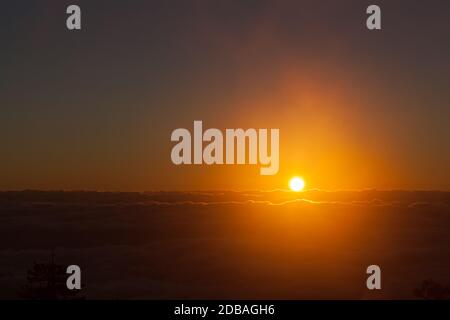 Die Sonne sieht aus wie ein leuchtender orangefarbener Feuerball Als es den Nebel und Nebel über einem erhellt Dicke Wolkenschicht, die das Tal am Sandia bedeckt Moun Stockfoto