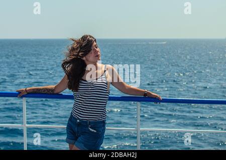 Hübsche junge Brunette-Frau in einem gestreiften Tanktop und blaue Denim-Shorts genießen das Meer auf dem Deck eines Schiffes Stockfoto