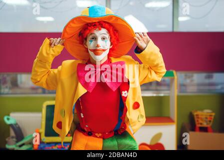 Lustige Clown mit Make-up in bunten Hut gekleidet und Kostüm Posen im Kinderbereich. Geburtstagsfeier im Spielzimmer, Babyurlaub auf dem Spielplatz. Kindisch Stockfoto