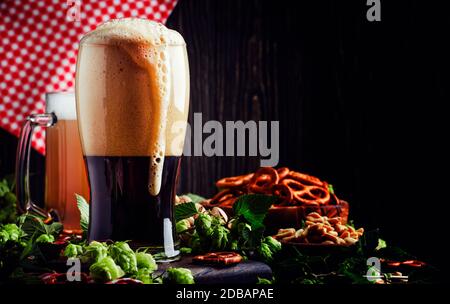 Dunkles deutsches Bier wird in ein Glas gegossen, frischer grüner Hopfen und Schalen mit salzigen Snacks und Nüssen, herbstliches Bierfest-Konzept, dunkler Hintergrund, Selecti Stockfoto