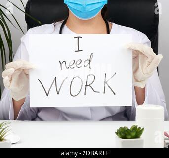 Frau Ärztin im weißen Mantel, sterile medizinische Handschuhe hält ein Plakat mit der Aufschrift Ich brauche Arbeit, Konzept der Arbeitslosigkeit und Jobsuche Stockfoto