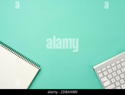 Weiße kabellose Aluminium-Tastatur und offenes Notebook mit leeren weißen Blättern auf grünem Hintergrund, Ansicht von oben, Arbeitsplatz Stockfoto