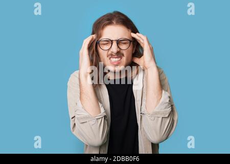 Bärtiger Kaukasusmann zeigt Kopfschmerzen, die seinen Kopf berühren Und schreiend an einer blauen Studiowand Stockfoto