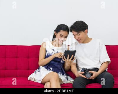 Asiatische Fotografen ermöglichen es Modellen, Bilder anzusehen, die auf dem Tablet-PC-Bildschirm aufgenommen wurden, während sie auf einem roten Sofa sitzen. Arbeitsatmosphäre im Fotostu Stockfoto
