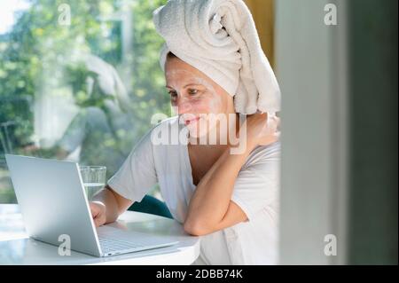 Frau mit Gesichtsmaske und Handtuch auf dem Kopf mit Laptop Am Tisch Stockfoto