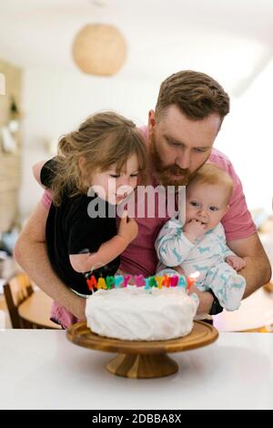 Vater trägt Kinder (2-3, 6-11 Monate) und bläst Geburtstagskerzen auf Kuchen Stockfoto
