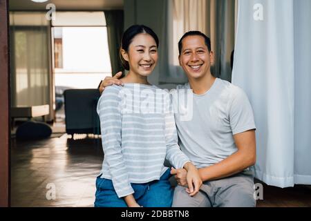 Portrait von jungen erwachsenen asiatischen Paar umarmt zusammen mit häuslichen Interieur im Hintergrund. 30s glücklich reifen Mann und Frau lächelnd und Blick auf die Kamera. Ehe und glückliche Beziehung Leben Konzept Stockfoto