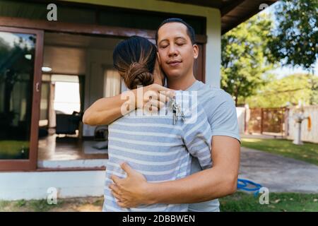 Portrait von 30s glücklich junge Erwachsene asiatische Paar umarmen, während ein Haus Schlüssel halten. Mann und Frau Immobilienbesitzer umarmen vor ihren Häusern. Konzept für die Umsetzung von Investitionen in den Wohnungsbau Stockfoto