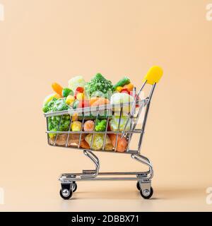 Gefrorenes Gemüse sortiert in Spielzeug Warenkorb auf cremefarbenem Hintergrund. Voll mit verschiedenen Tiefkühlgemüse Food Shop Trolley auf beige oder gelbe Hintergrund. Stockfoto