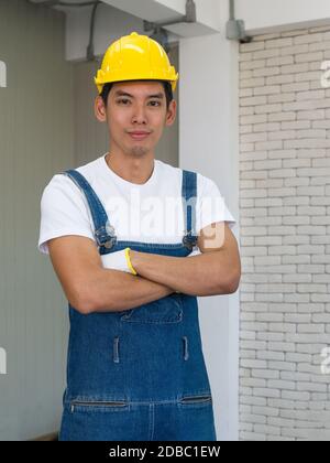 Ein asiatischer Zimmermann, der einen gelben Hut trägt, stellte sich im Werkstattzimmer mit den Armen. Stockfoto
