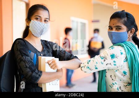 Konzept der Hochschule wieder öffnen, zwei Mädchen in medizinischer Maske Gruß mit Ellenbogen Beule mit Studenten im Hintergrund während Coronavirus oder covid-19 Pandemie Stockfoto
