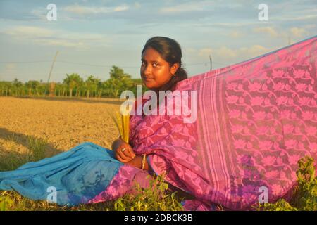 Nahaufnahme eines Teenagers mit rosa Sari, Ohrringen, Halskette, mit langen dunklen Haaren auf einem landwirtschaftlichen Feld sitzend mit ihrem Saree gestreckt Stockfoto