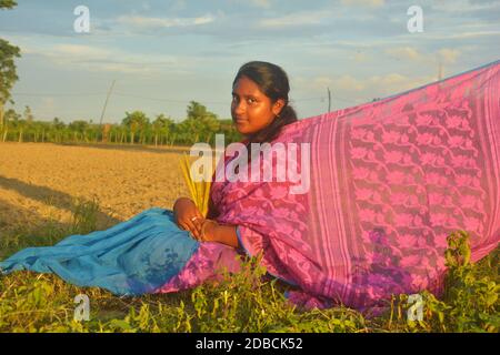 Nahaufnahme eines Teenagers mit rosa Sari, Ohrringen, Halskette, mit langen dunklen Haaren auf einem landwirtschaftlichen Feld sitzend mit ihrem Saree gestreckt Stockfoto