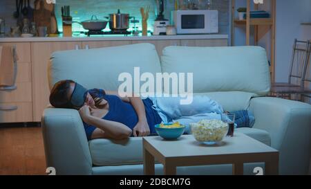 Frau schläft mit Schlafmaske im Wohnzimmer während tv-Show. Müde erschöpft einsam schläfrig Dame im Schlafanzug Einschlafen auf gemütlichen Sofa vor dem Fernseher, die Augen schließen, während Film in der Nacht zu sehen. Stockfoto