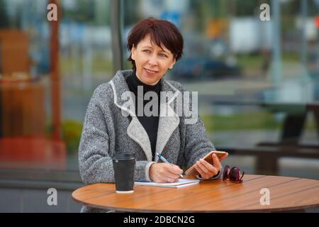 Eine Geschäftsfrau sitzt an einem Tisch in einem Sommercafé. Eine Frau hält ein Smartphone in der Hand und schreibt in ein Notebook. Stockfoto