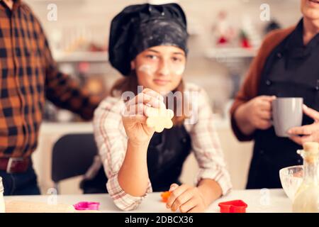 Enkelkind am weihnachtstag hält geformte Pastelle trägt Schürze und bonette. Happy fröhlich freudig Teenager-Mädchen hilft älteren Frau Vorbereitung süße Kekse, um Winterferien zu feiern. Stockfoto