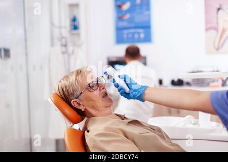 Zahnarzthelferin mit digitaler Körpertemperaturanzeige vor dem auf dem Stuhl sitzenden Patienten. Medizinische Facharzt in der Zahnklinik unter Patienten Temperatur mit digitalen Gerät. Stockfoto