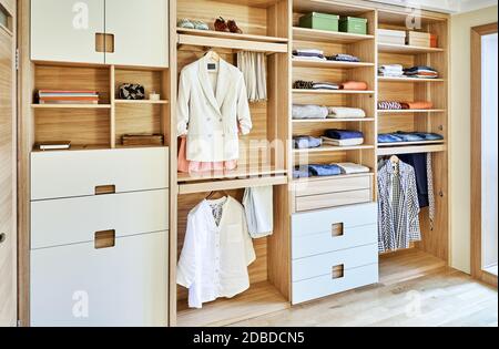 Interne Details der Holzgarderobe mit ausziehbarem Gestell für Kleiderständer. Moderner Kleiderschrank mit Kleidung, die auf ausziehbaren Racks hängt und auf dem gefaltet ist Stockfoto