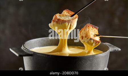 Im Winter wird ein köstliches Schmelzkäse-Fondue in einem gusseisernen Topf serviert, mit Gabeln, die geröstete Baguette und aufsteigenden Dampf auf dunklem Hintergrund eintauchen Stockfoto