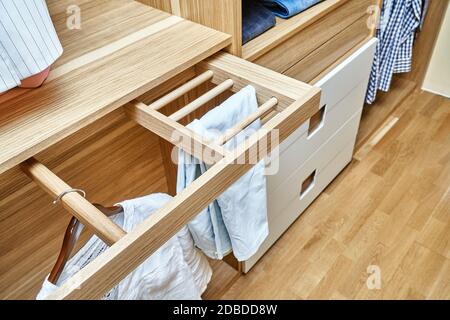 Interne Details der Holzgarderobe mit ausziehbarem Gestell für Kleiderständer. Moderner Kleiderschrank mit Kleidung, die auf ausziehbaren Racks hängt und auf dem gefaltet ist Stockfoto