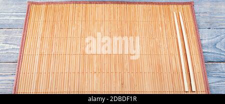 Zwei hölzerne Essstäbchen liegen auf dem Rand einer Bambusmatte, die auf einem Holztisch aus strukturierten Brettern sitzt. Stockfoto