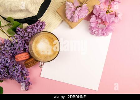 Leeres Papier auf Zwischenablage, Sommermock-up. Ein leeres Blatt Papier, umgeben von einem Strandhut, rosa Blumen, Kaffee und Makronen, steht auf einem rosa Hintergrund Stockfoto