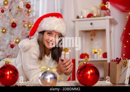 Happy Girl im Weihnachtskleid liegt auf dem Boden in der Nähe des Weihnachtsbaums mit einem Geschenk Stockfoto