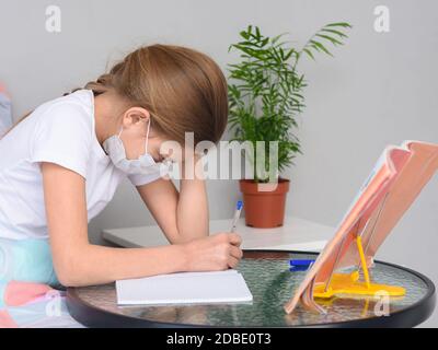 Ein Mädchen in einer medizinischen Maske macht Hausaufgaben an einem Tisch vor dem Bett Stockfoto