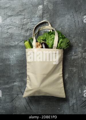 Baumwolltasche mit Gemüse auf grauem Hintergrund. Bio-Baumwoll-Canvas-Tasche mit frischen Zucchini, Auberginen, Ingwerwurzel, Brokkoli und Salat. Vertikal. Mock up für Design oder Ihr Logo Stockfoto