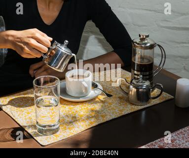 Die Hand einer Frau gießen frische Milch aus dem Topf in eine Kaffeetasse, mit einem französischen Presse und Metall Service in einem Restaurant. Stockfoto